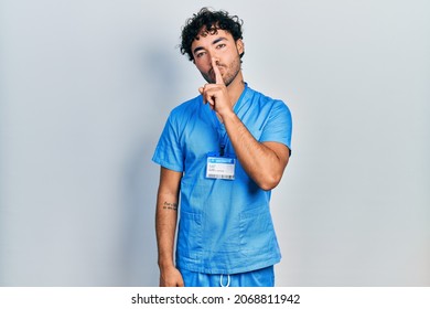 Young hispanic man wearing blue male nurse uniform asking to be quiet with finger on lips. silence and secret concept. 