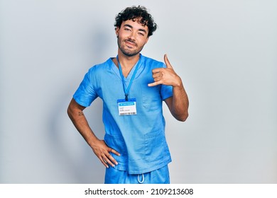 Young Hispanic Man Wearing Blue Male Nurse Uniform Smiling Doing Phone Gesture With Hand And Fingers Like Talking On The Telephone. Communicating Concepts. 