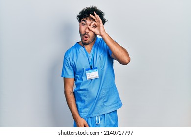 Young Hispanic Man Wearing Blue Male Nurse Uniform Doing Ok Gesture Shocked With Surprised Face, Eye Looking Through Fingers. Unbelieving Expression. 