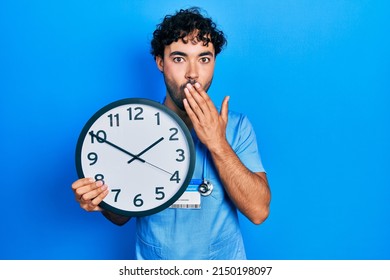 Young Hispanic Man Wearing Blue Male Nurse Uniform Holding Clock Covering Mouth With Hand, Shocked And Afraid For Mistake. Surprised Expression 