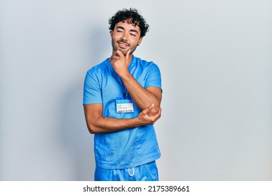 Young Hispanic Man Wearing Blue Male Nurse Uniform With Hand On Chin Thinking About Question, Pensive Expression. Smiling With Thoughtful Face. Doubt Concept. 