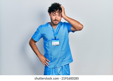 Young Hispanic Man Wearing Blue Male Nurse Uniform Confuse And Wonder About Question. Uncertain With Doubt, Thinking With Hand On Head. Pensive Concept. 