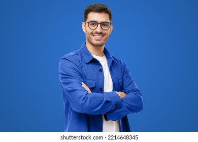 Joven hispano con camisa azul y gafas, mirando la cámara con una sonrisa positiva y segura de sí mismo, con los brazos cruzados, aislado de fondo azul