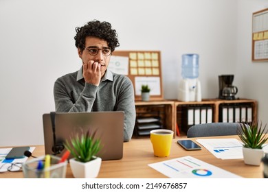 Young hispanic man wearing business style sitting on desk at office looking stressed and nervous with hands on mouth biting nails. anxiety problem. 