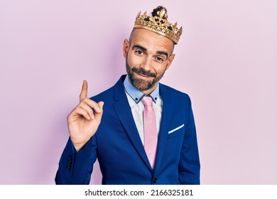 Young Hispanic Man Wearing Business Clothes And King Crown Smiling With An Idea Or Question Pointing Finger Up With Happy Face, Number One 