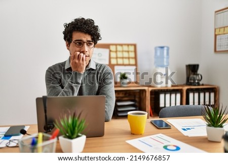 Young hispanic man wearing business style sitting on desk at office looking stressed and nervous with hands on mouth biting nails. anxiety problem. 