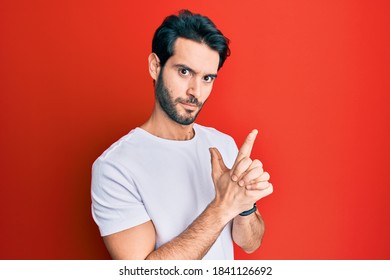 Young Hispanic Man Wearing Casual White Tshirt Holding Symbolic Gun With Hand Gesture, Playing Killing Shooting Weapons, Angry Face 