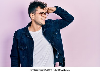 Young hispanic man wearing casual clothes and glasses very happy and smiling looking far away with hand over head. searching concept. 