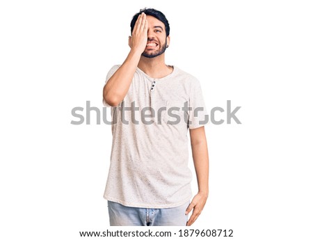 Young hispanic man wearing casual clothes covering one eye with hand, confident smile on face and surprise emotion. 