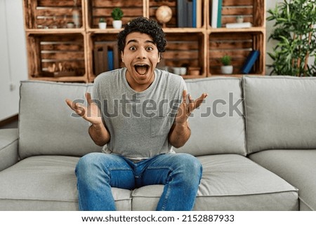 Young hispanic man wearing casual clothes sitting on the sofa at home celebrating victory with happy smile and winner expression with raised hands 