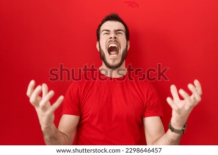 Young hispanic man wearing casual red t shirt crazy and mad shouting and yelling with aggressive expression and arms raised. frustration concept. 