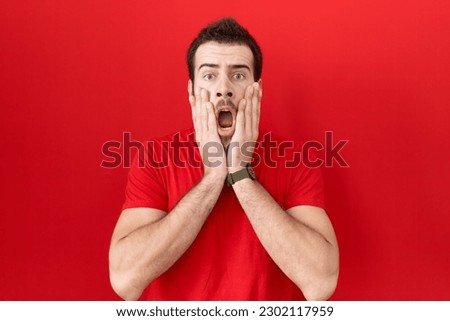 Young hispanic man wearing casual red t shirt afraid and shocked, surprise and amazed expression with hands on face 