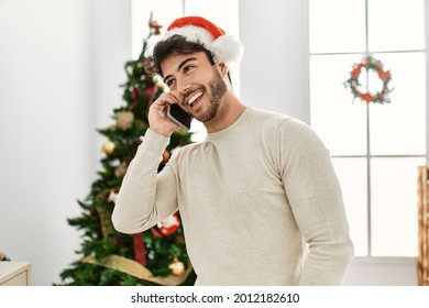 Young Hispanic Man Wearing Christmas Hat Talking On The Smartphone At Home.