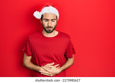 Young Hispanic Man Wearing Christmas Hat With Hand On Stomach Because Indigestion, Painful Illness Feeling Unwell. Ache Concept. 