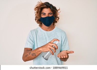 Young Hispanic Man Wearing Covid-19 Mask Using Hand Sanitizer Gel Winking Looking At The Camera With Sexy Expression, Cheerful And Happy Face. 