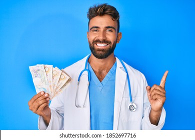Young hispanic man wearing doctor uniform holding uk pounds banknotes smiling happy pointing with hand and finger to the side 