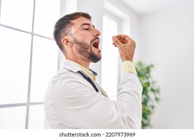 Young Hispanic Man Wearing Doctor Uniform Taking Pill At Clinic