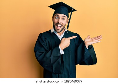 Young hispanic man wearing graduation cap and ceremony robe amazed and smiling to the camera while presenting with hand and pointing with finger. 