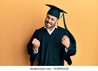 Young hispanic man wearing graduation cap and ceremony robe very happy and excited doing winner gesture with arms raised, smiling and screaming for success. celebration concept. 