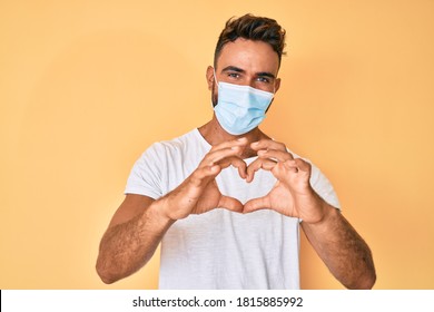 Young Hispanic Man Wearing Medical Mask Smiling In Love Doing Heart Symbol Shape With Hands. Romantic Concept. 