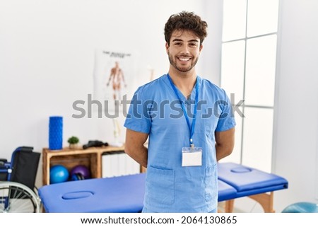 Young hispanic man wearing physio therapist uniform standing at clinic
