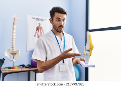 Young Hispanic Man Wearing Physiotherapist Uniform Holding Anatomical Model Of Knee At Rehab Clinic