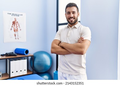 Young hispanic man wearing physiotherapist uniform standing with arms crossed gesture at clinic - Shutterstock ID 2143152351