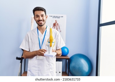 Young Hispanic Man Wearing Physiotherapist Uniform Holding Anatomical Model Of Knee At Rehab Clinic