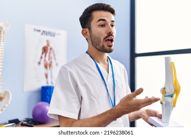 Young Hispanic Man Wearing Physiotherapist Uniform Holding Anatomical Model Of Knee At Rehab Clinic