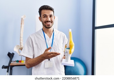 Young Hispanic Man Wearing Physiotherapist Uniform Holding Anatomical Model Of Knee At Rehab Clinic