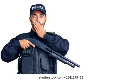 Young Hispanic Man Wearing Police Uniform Holding Shotgun Covering Mouth With Hand, Shocked And Afraid For Mistake. Surprised Expression 