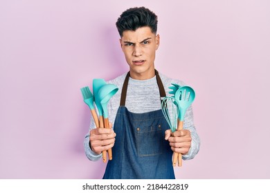 Young Hispanic Man Wearing Professional Baker Apron Holding Cooking Tools In Shock Face, Looking Skeptical And Sarcastic, Surprised With Open Mouth 