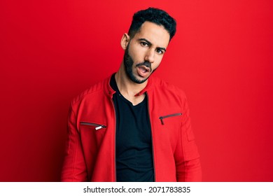 Young Hispanic Man Wearing Red Leather Jacket In Shock Face, Looking Skeptical And Sarcastic, Surprised With Open Mouth 