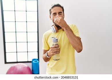 Young Hispanic Man Wearing Sportswear And Drinking Water At The Gym Smelling Something Stinky And Disgusting, Intolerable Smell, Holding Breath With Fingers On Nose. Bad Smell 