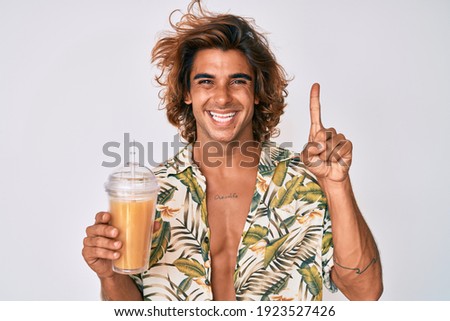 Young hispanic man wearing summer shirt drinking glass of orange juice smiling with an idea or question pointing finger with happy face, number one 