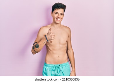 Young hispanic man wearing swimwear shirtless doing happy thumbs up gesture with hand. approving expression looking at the camera showing success. 