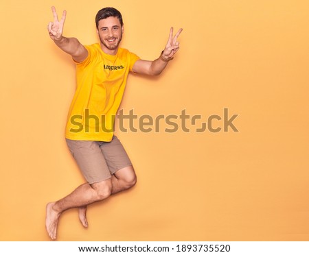 Young hispanic man wearing t shirt with happiness word message smiling happy. Jumping with smile on face doing victory sign over isolated yellow background