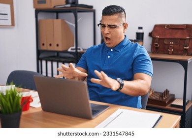 Young hispanic man working at the office with laptop afraid and terrified with fear expression stop gesture with hands, shouting in shock. panic concept. 