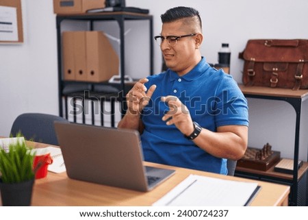 Young hispanic man working at the office with laptop disgusted expression, displeased and fearful doing disgust face because aversion reaction. with hands raised 