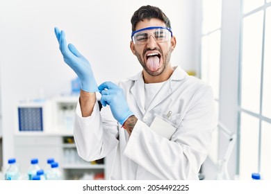 Young Hispanic Man Working At Scientist Laboratory Putting Gloves On Sticking Tongue Out Happy With Funny Expression. 