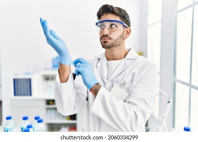 Young Hispanic Man Working At Scientist Laboratory Putting Gloves On Smiling Looking To The Side And Staring Away Thinking. 