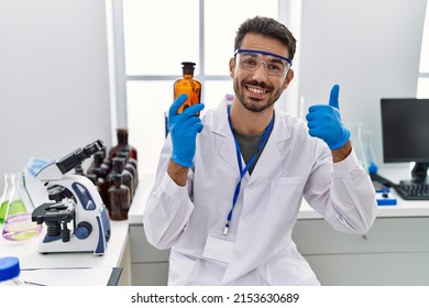 Young Hispanic Man Working At Scientist Laboratory Holding Bottle Smiling Happy And Positive, Thumb Up Doing Excellent And Approval Sign 