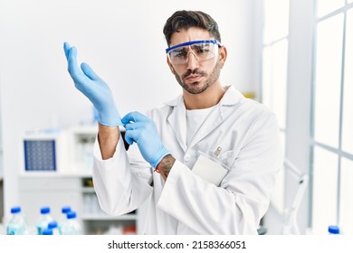 Young Hispanic Man Working At Scientist Laboratory Putting Gloves On Skeptic And Nervous, Frowning Upset Because Of Problem. Negative Person. 