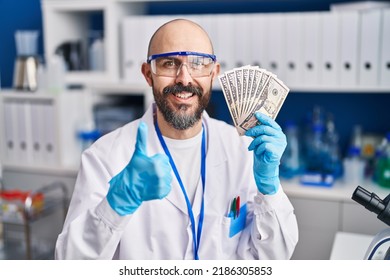 Young Hispanic Man Working At Scientist Laboratory Holding Money Smiling Happy And Positive, Thumb Up Doing Excellent And Approval Sign 