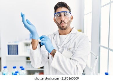 Young Hispanic Man Working At Scientist Laboratory Putting Gloves On Making Fish Face With Mouth And Squinting Eyes, Crazy And Comical. 