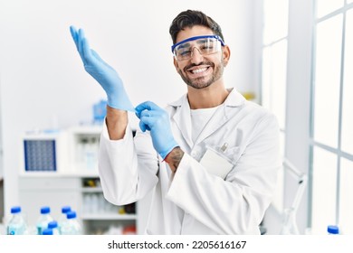 Young Hispanic Man Working At Scientist Laboratory Putting Gloves On Smiling With A Happy And Cool Smile On Face. Showing Teeth. 