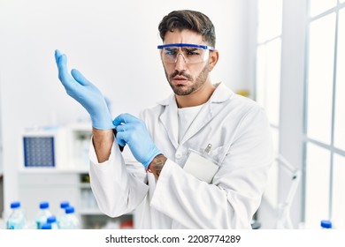 Young Hispanic Man Working At Scientist Laboratory Putting Gloves On Clueless And Confused Expression. Doubt Concept. 