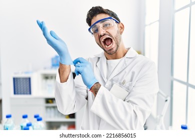 Young Hispanic Man Working At Scientist Laboratory Putting Gloves On Celebrating And Amazed For Success With Open Eyes Screaming Excited. 