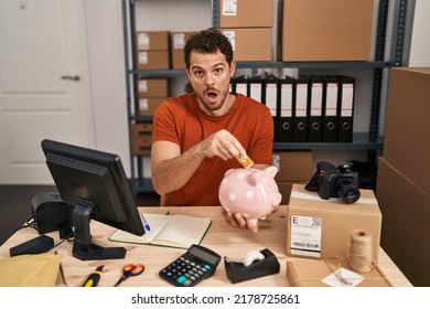 Young Hispanic Man Working At Small Business Ecommerce Holding Piggy Bank In Shock Face, Looking Skeptical And Sarcastic, Surprised With Open Mouth 