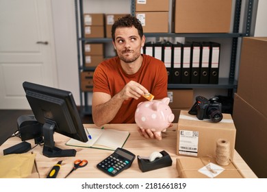 Young Hispanic Man Working At Small Business Ecommerce Holding Piggy Bank Smiling Looking To The Side And Staring Away Thinking. 